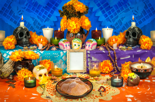Altar for Dia de los Muertos with sugar skulls, marigolds, bread of the dead, candles and blue, purple and orange paper decorations.