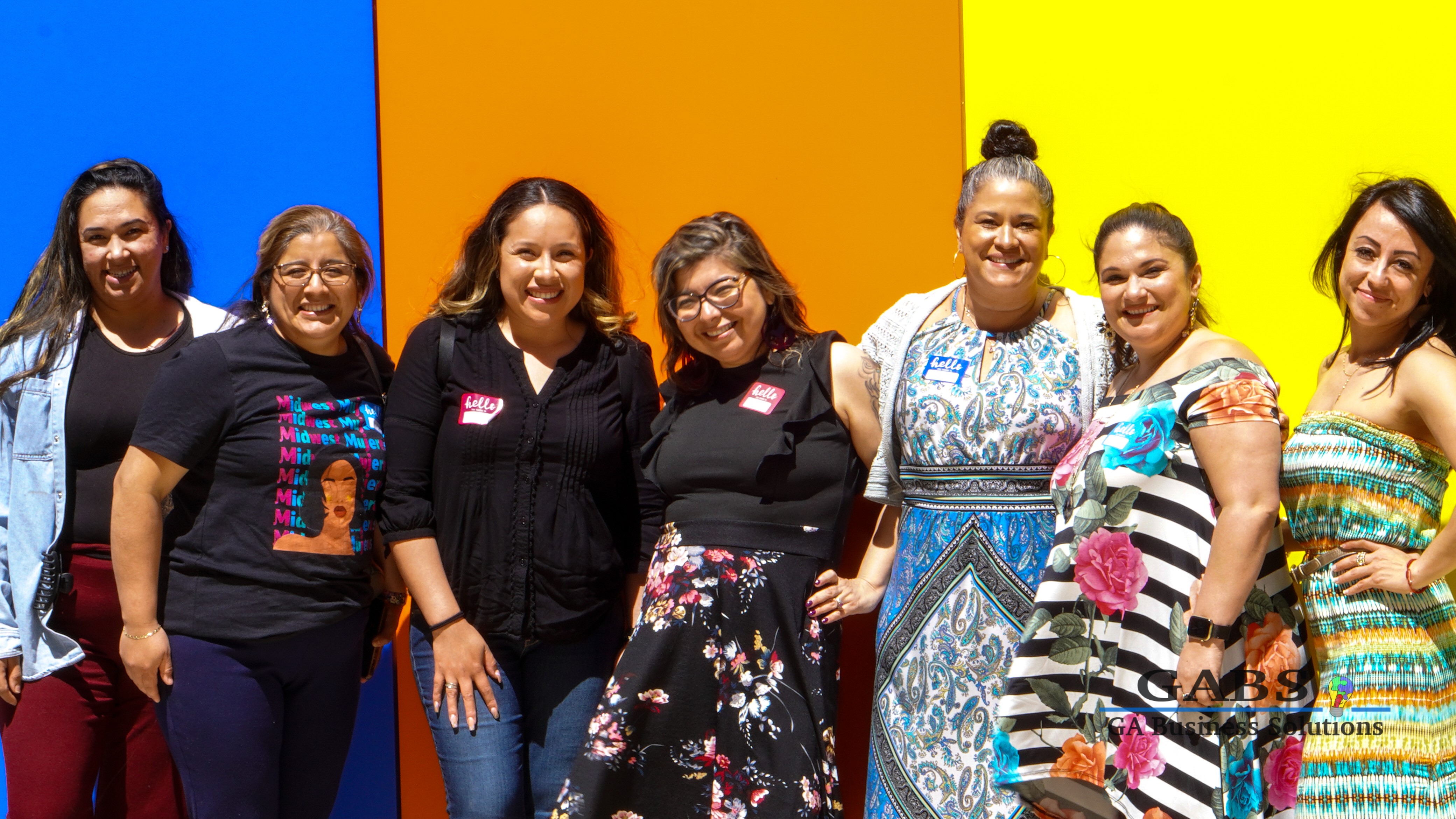 Araceli Esparza (center) with 6 other Latina women standing in front of a bright blue, orange and yellow backdrop and smiling.