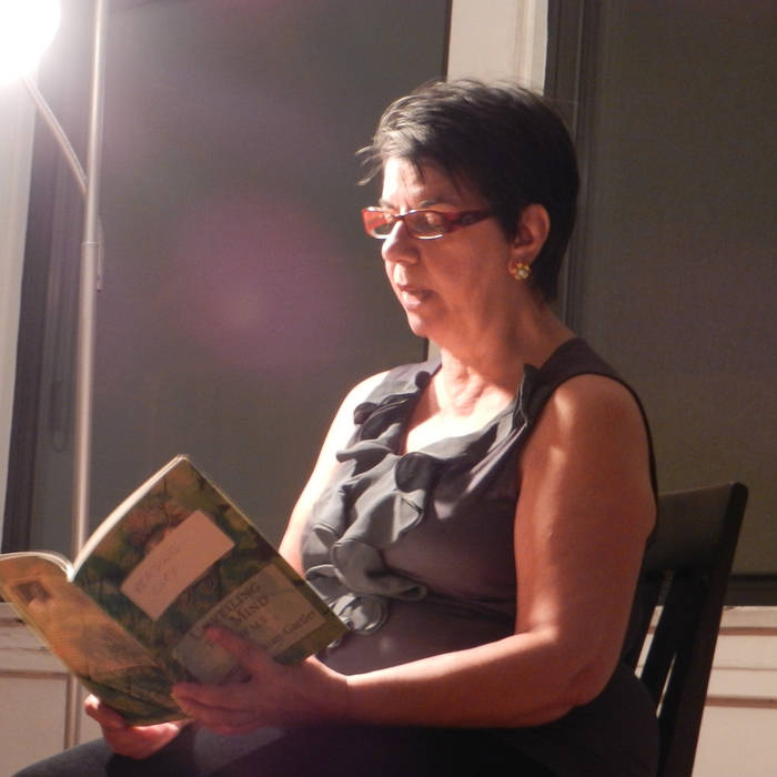 Beatriz Badikian-Gartler reading from a book of poetry against a dark background with a light shining down on her face.