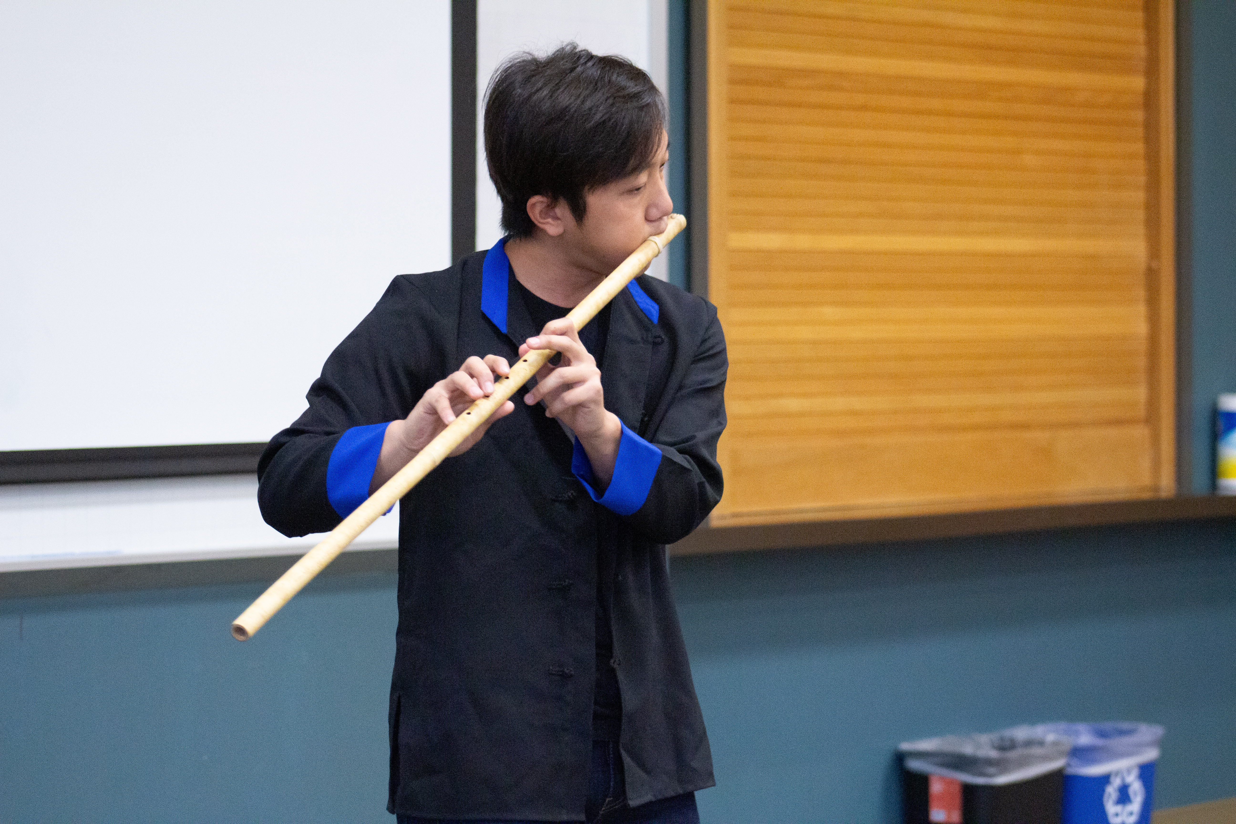 Neng Now, a HMong man with short hair wearing a black jacket with blue cuffs plays  a traditional wooden flute.