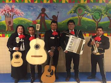 Mariachi Sol stands in front of a mural. There are five members, each holding various instruments.