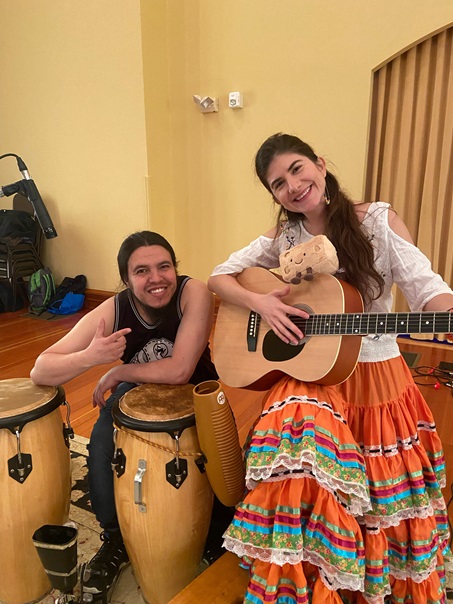 A man sits behind two drums, Angela Puerta sits next to him holding a guitar