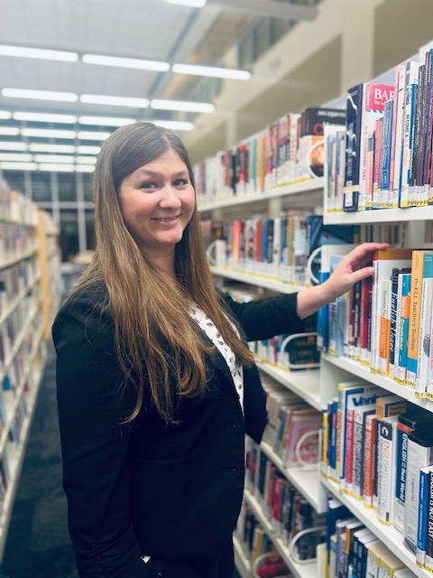 Michelle Gibbons Michelle Gibbons, a white woman with long light brown hair, stands smiling in the aisle of a library and reaches for a book on a shelf.
