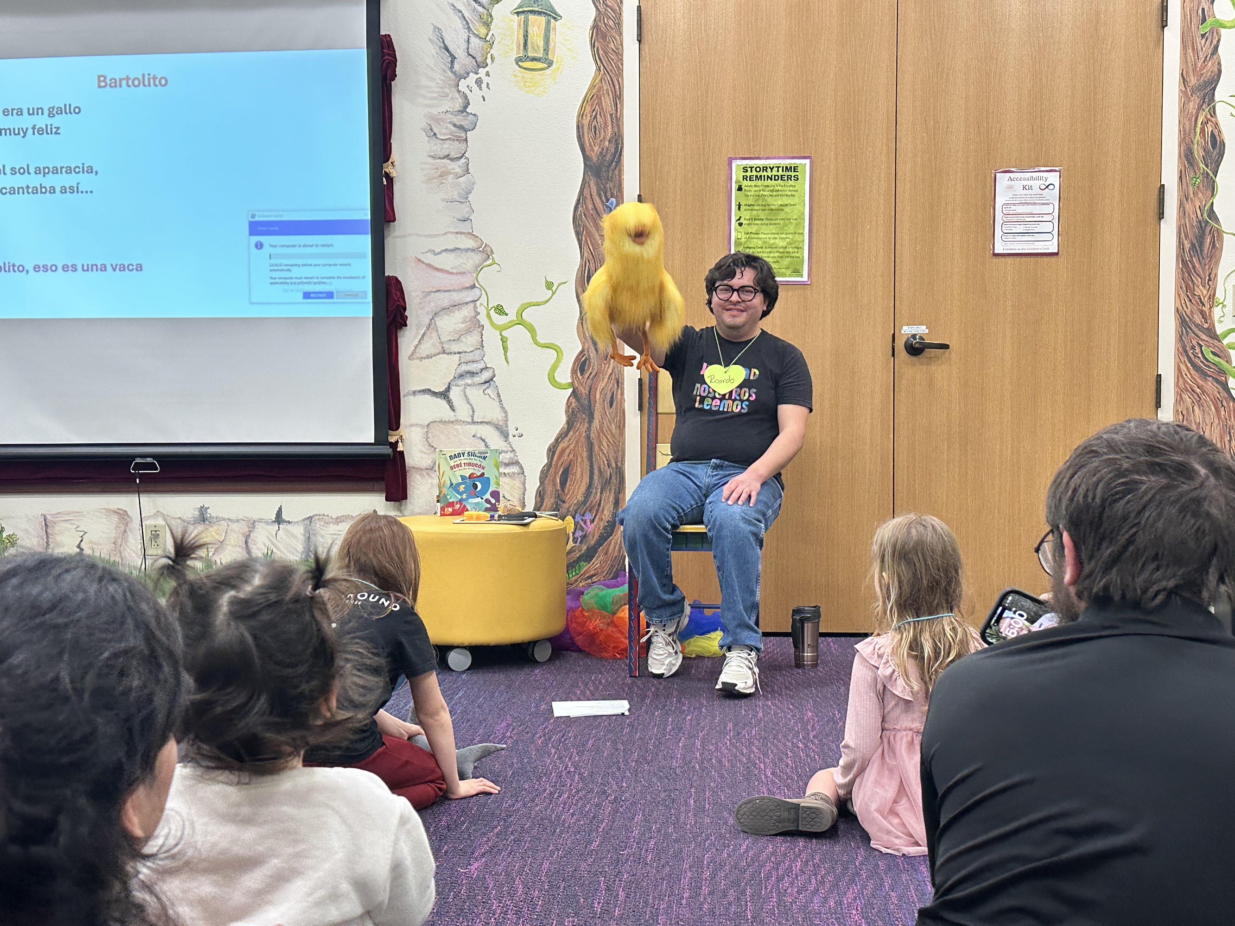 Spanish storytime with Ricardo Marroquin Ricardo Marroquin smiling and holding a large, yellow bird puppet while singing to a group of young kids at Sun Prairie Public Library