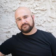 A middle-aged white man with a bald head and full, dark beard wearing a black t-shirt.